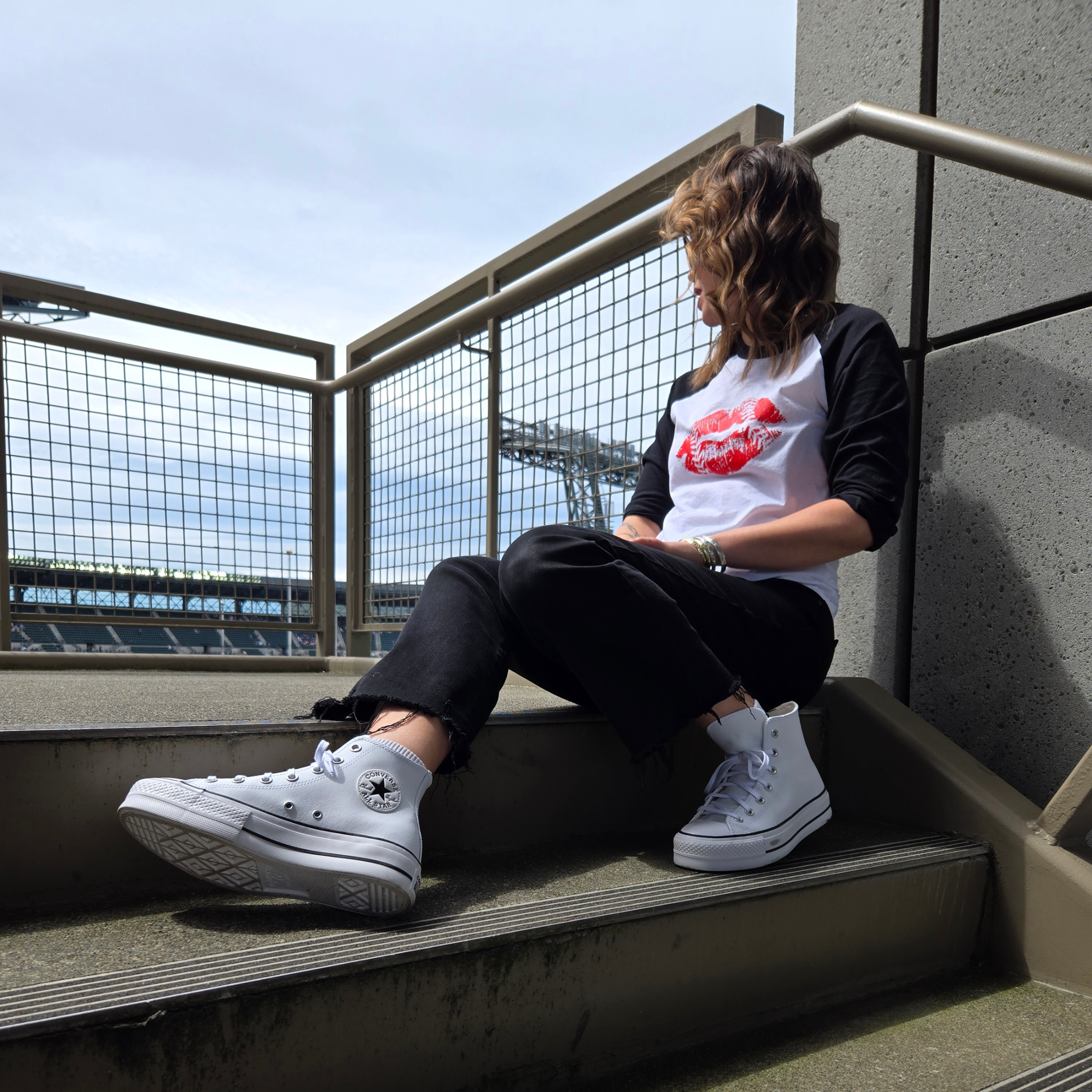 A woman sits on outdoor concrete stairs wearing a black and white raglan t-shirt with a red lip stick graphic, black jeans, and white high-top sneakers with stadium in the background.
