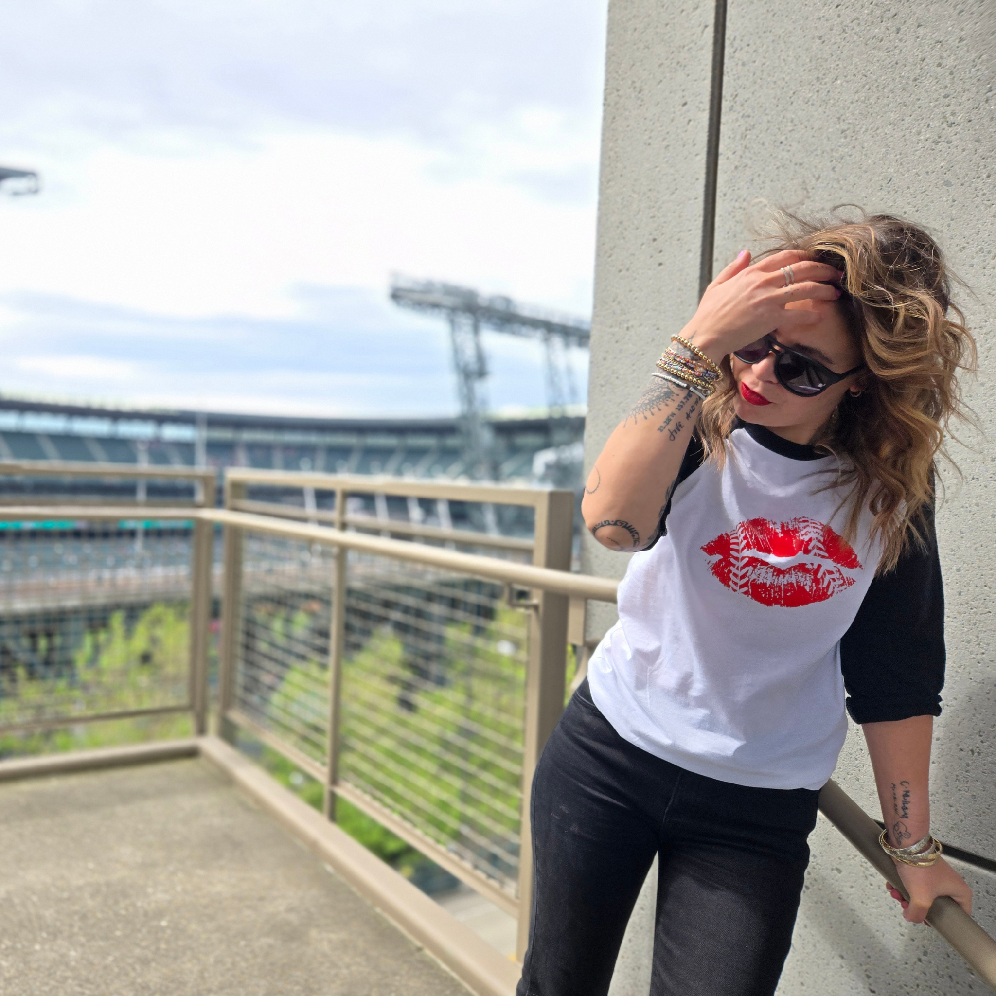 A woman posing on an outdoor balcony wearing a white and black raglan t-shirt with a red lips graphic, black jeans, and sunglasses, with a stadium in the background.