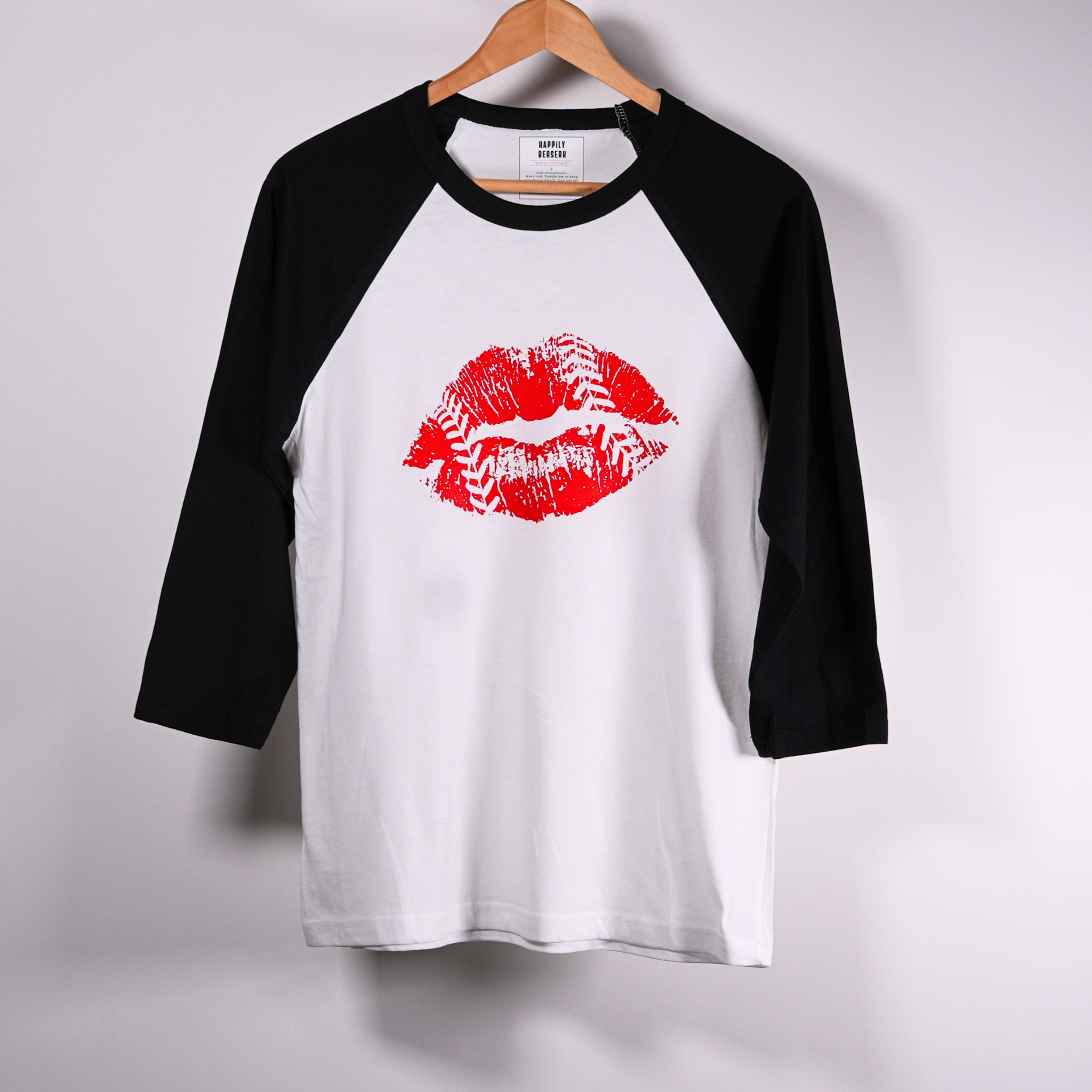 A 3/4-sleeve raglan baseball t-shirt hanging on a  hanger against a light  wall. The shirt has a white body, black sleeves, and a red lipstick kiss-print graphic on the chest.