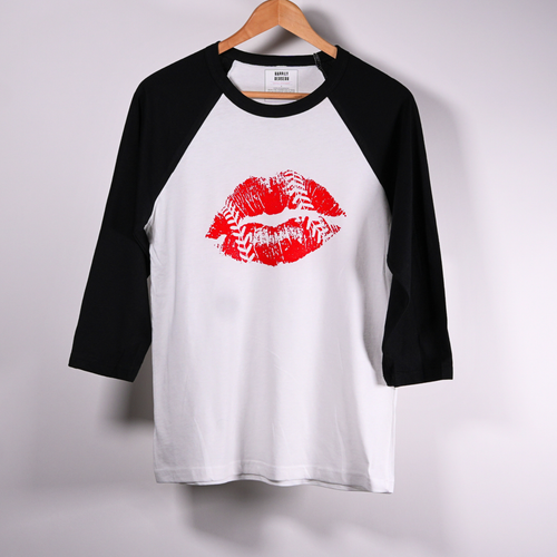 A 3/4-sleeve raglan baseball t-shirt hanging on a  hanger against a light  wall. The shirt has a white body, black sleeves, and a red lipstick kiss-print graphic on the chest.
