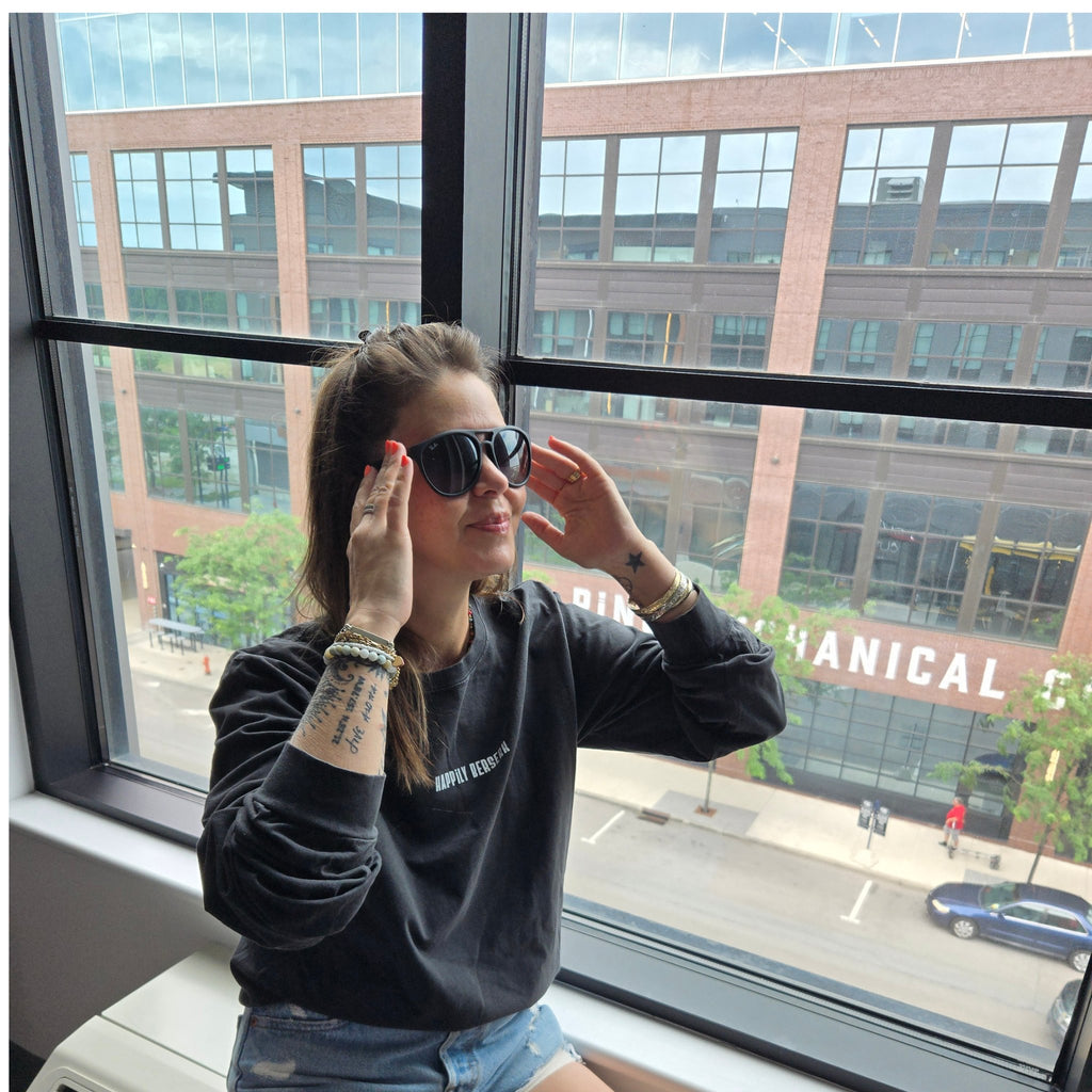A woman wearing a vintage black long-sleeve t-shirt with happily berserk in vintage white and denim shorts sits by a large window, adjusting black sunglasses.