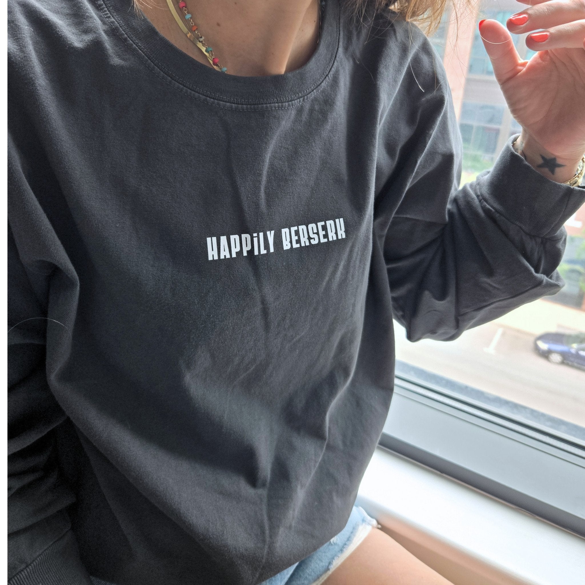A person wearing vintage black long-sleeve t-shirt with the text 'HAPPILY berserk' printed in vintage white across the chest, sitting by a window.