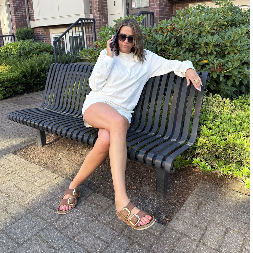 A woman wearing a vintage white long-sleeve t-shirt with 'happily berserk' printed on the chest with matching vintage white happily berserk shorts with side pockets sitting on a bench
