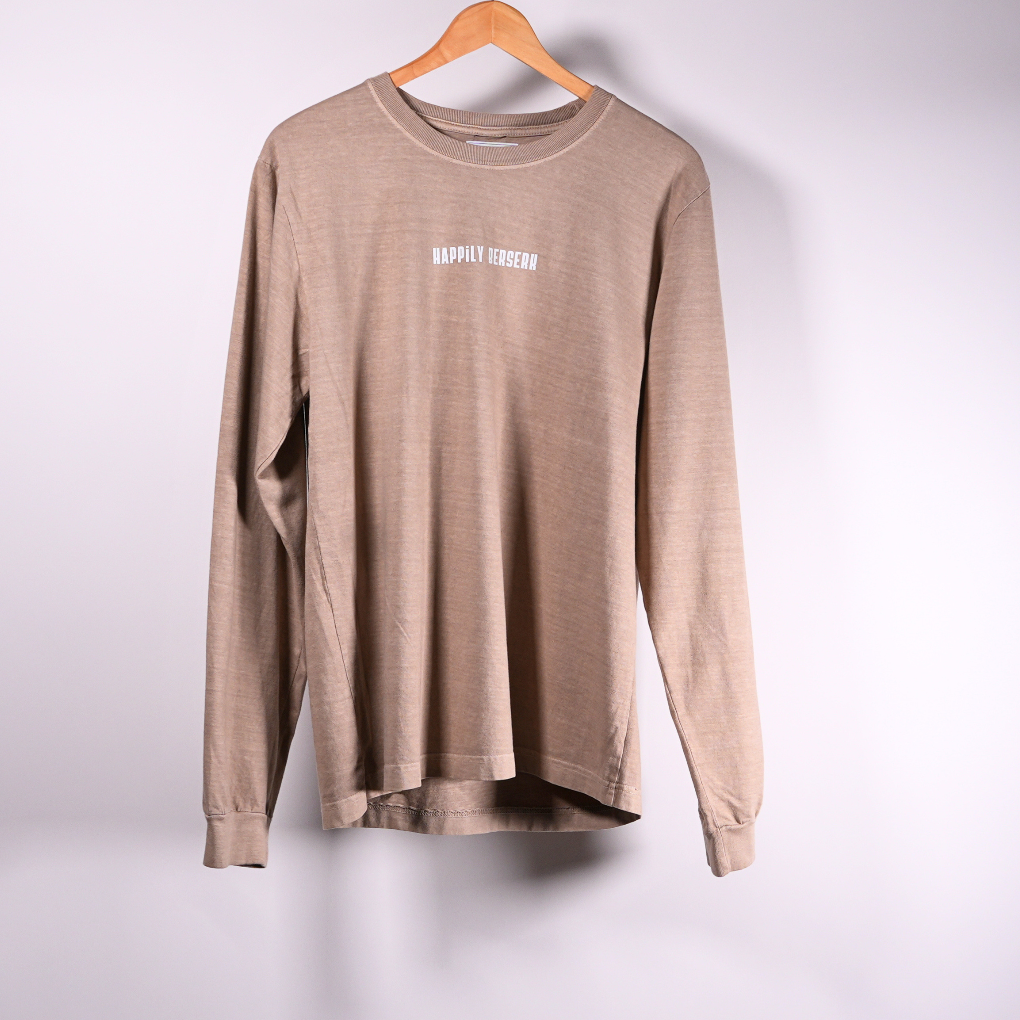 A vintage tan crewneck shirt hanging on a wooden hanger against a light  background, featuring the text 'HAPPILY BERSERK' printed in VINTAGE white on the chest.