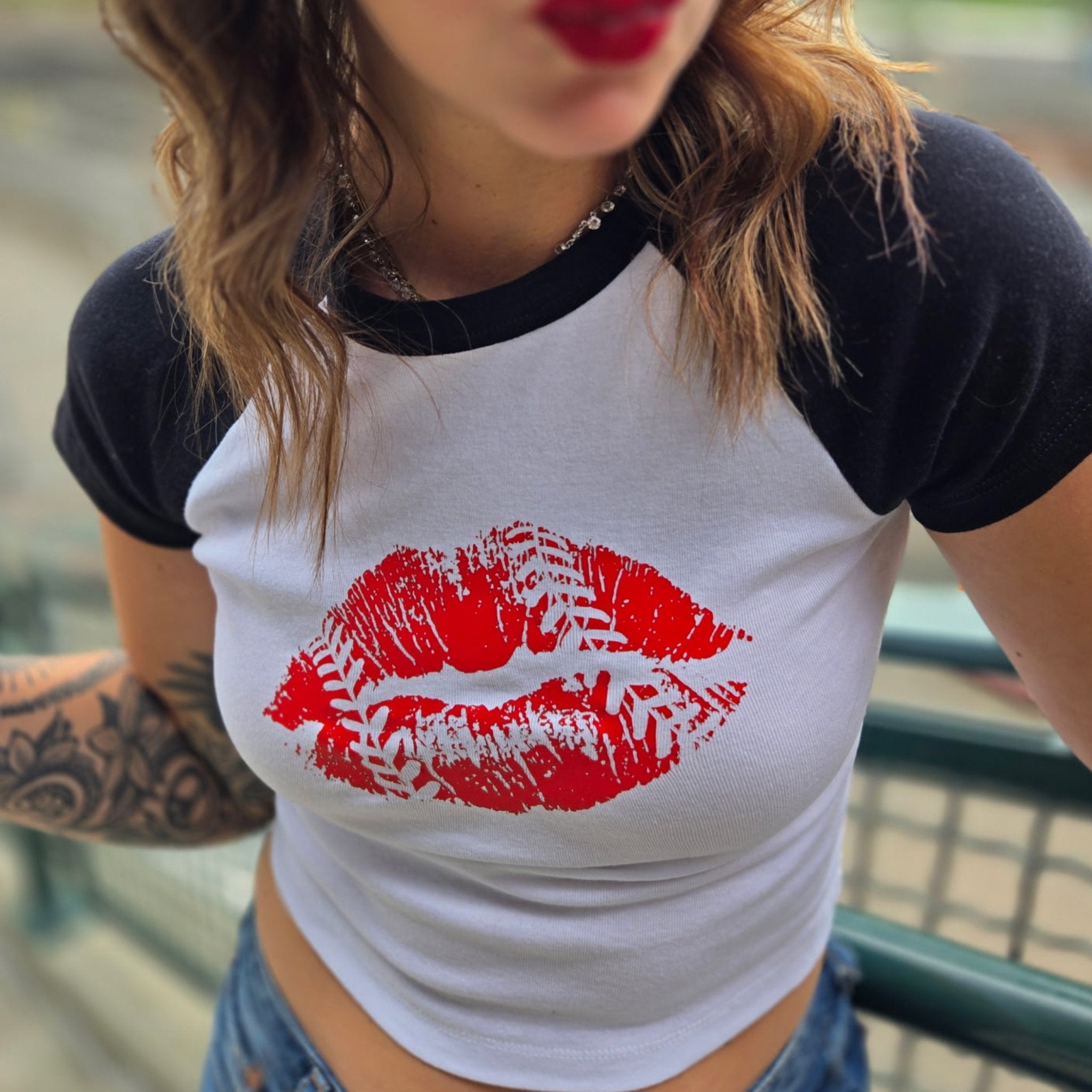 A close-up of a person wearing a white and black raglan baby baseball t-shirt with a large red lipstick kiss and baseball stitching print on the front, paired with blue jeans.