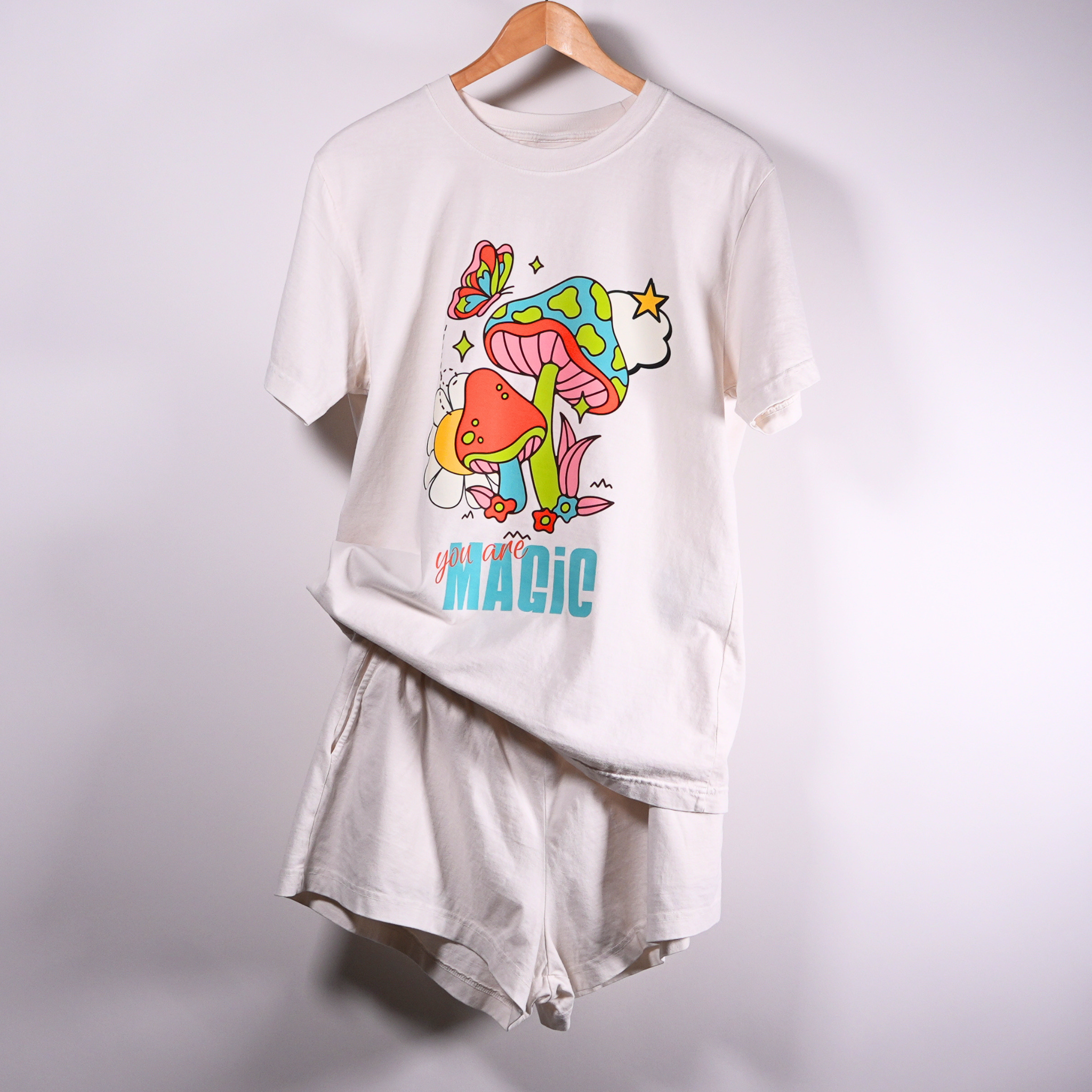 A vintage white short-sleeve t-shirt hanging on a wooden hanger against a light wall. The shirt features a colorful graphic print of a mushroom, flower, and butterfly with the text 'you are MAGIC' printed below with matching vintage white happily berserk shorts