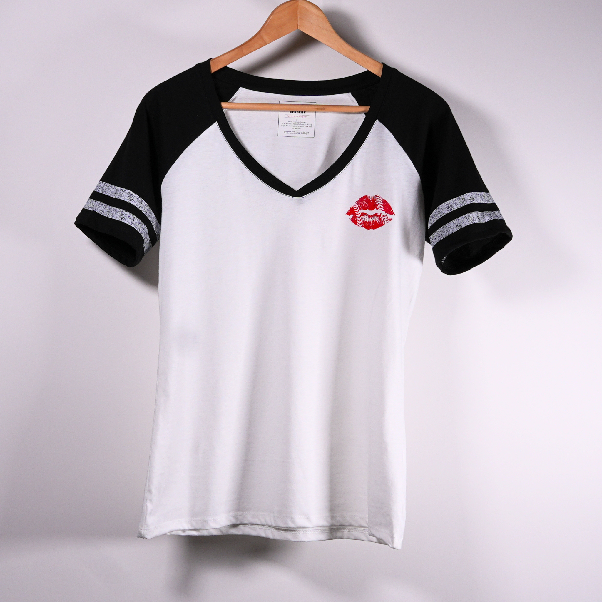 A short-sleeve V-Neck baseball t-shirt hanging on a wooden hanger. The shirt is white on the body with black sleeves and a small red lip graphic on the left chest.