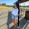 A woman wearing a white and black baseball t-shirt with a red lip logo on the chest, paired with denim shorts and brown sandals, standing outdoors next to a black pole at a baseball field