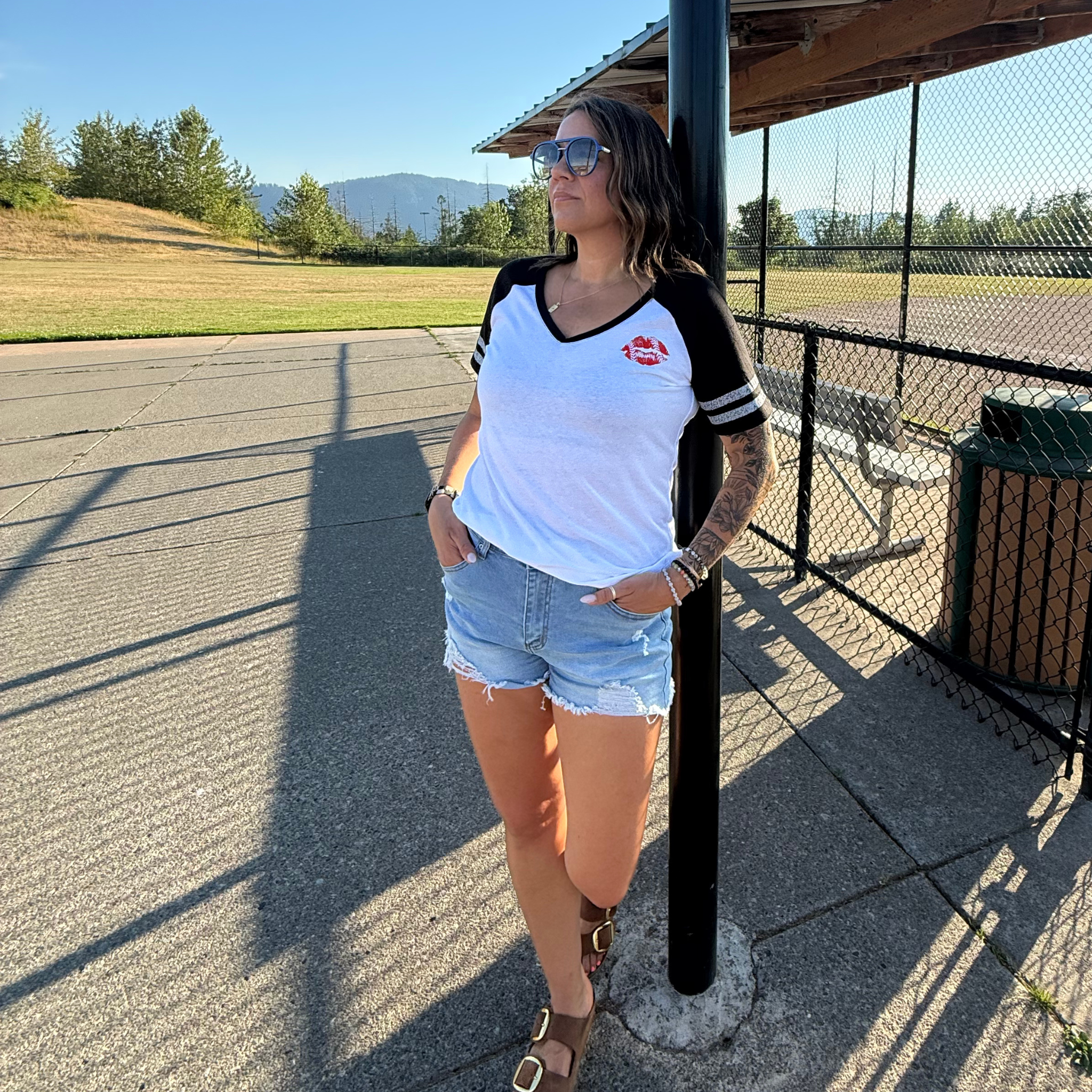 A woman wearing a white and black baseball t-shirt with a red lip logo on the chest, paired with denim shorts and brown sandals, standing outdoors next to a black pole at a baseball field
