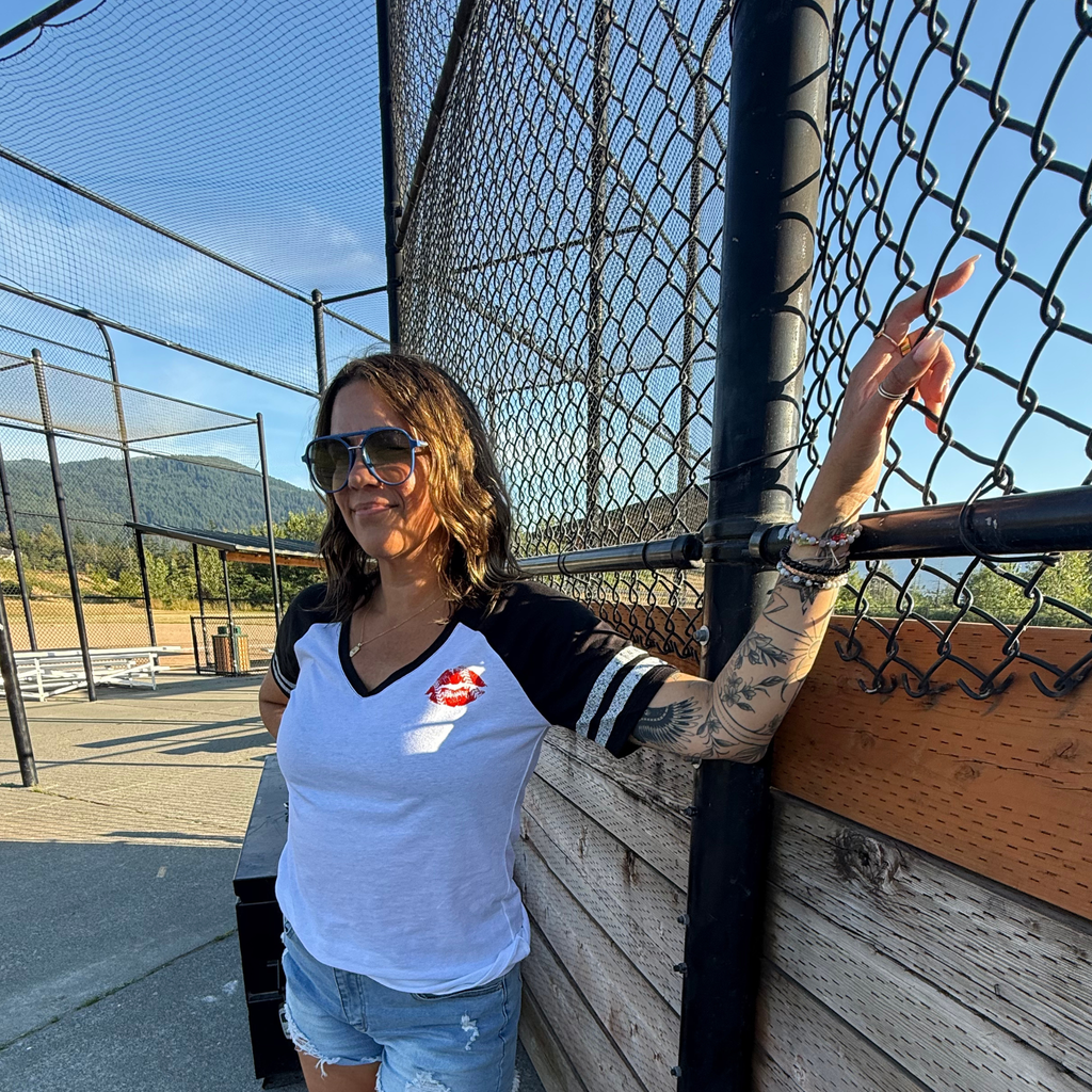 A woman wearing a white and black baseball t-shirt with a red lips graphic and sunglasses, standing next to a chain-link fence at an outdoor baseball field.