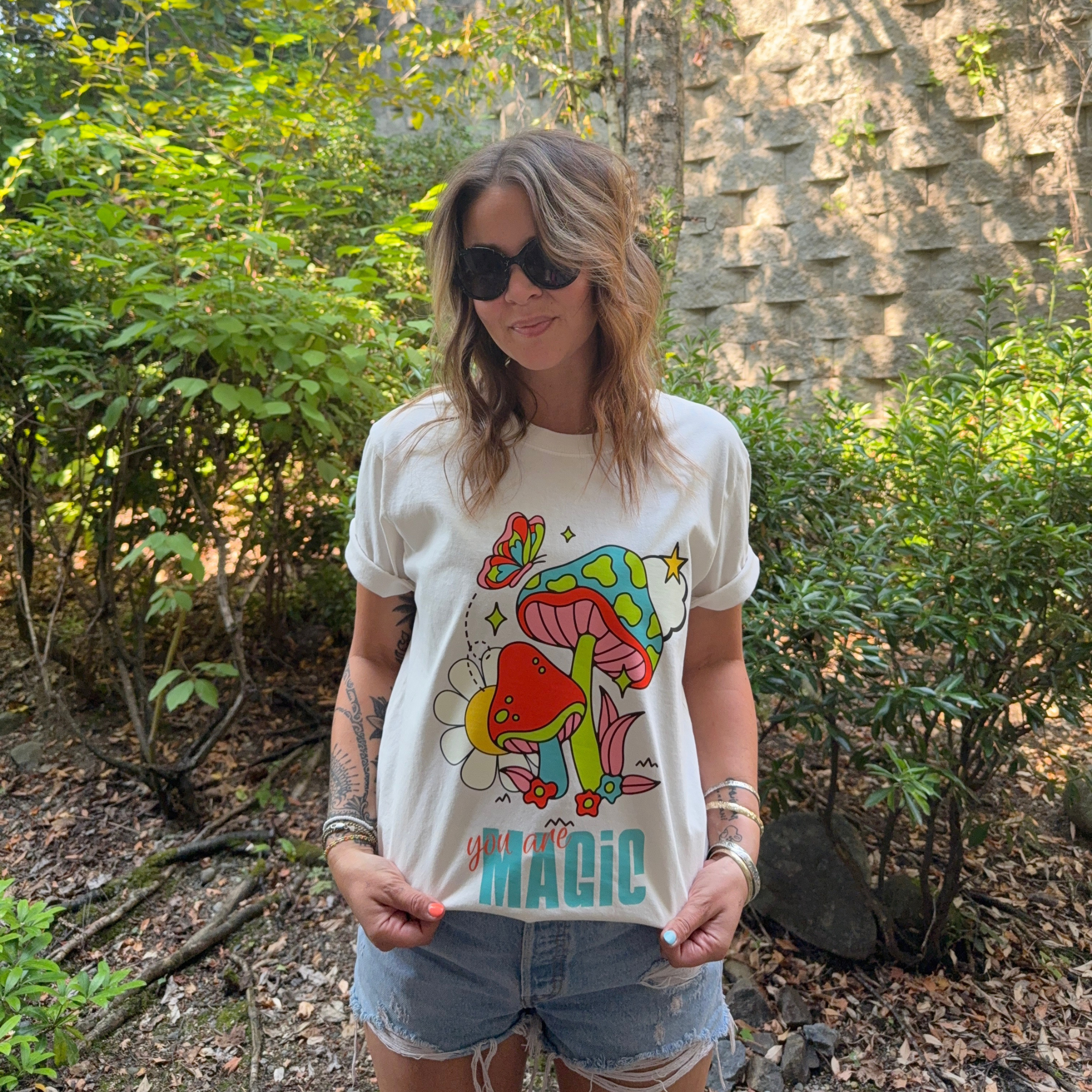 A woman wearing a a vintage white short-sleeve t-shirt hanging on a wooden hanger against a light wall. The shirt features a colorful graphic print of a mushroom, flower, and butterfly with the text 'you are MAGIC' printed below paired with denim shorts, standing outdoors in a garden setting.