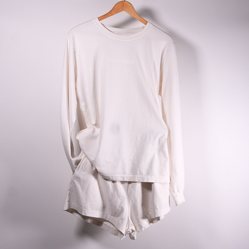 A white long-sleeve happily berserk crewneck long sleeve t-shirt and matching shorts set in A long-sleeve happily berserk crewneck t-shirt in vintage white, hanging on a wooden hanger against a plain light  wall.