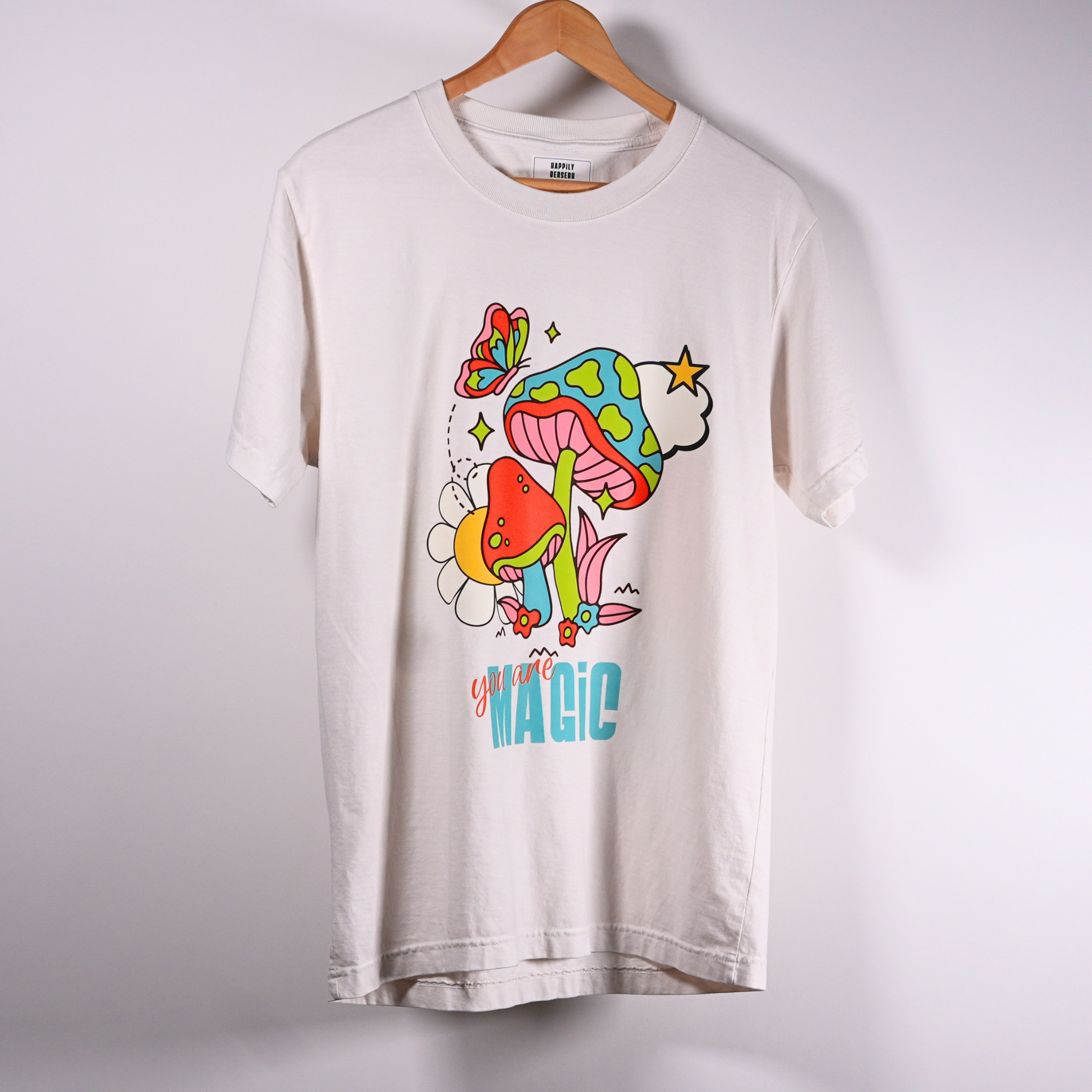 A vintage white short-sleeve t-shirt hanging on a wooden hanger against a light wall. The shirt features a colorful graphic print of a mushroom, flower, and butterfly with the text 'you are MAGIC' printed below.